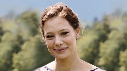 Person with hair pulled back standing outside in front of trees and blue sky