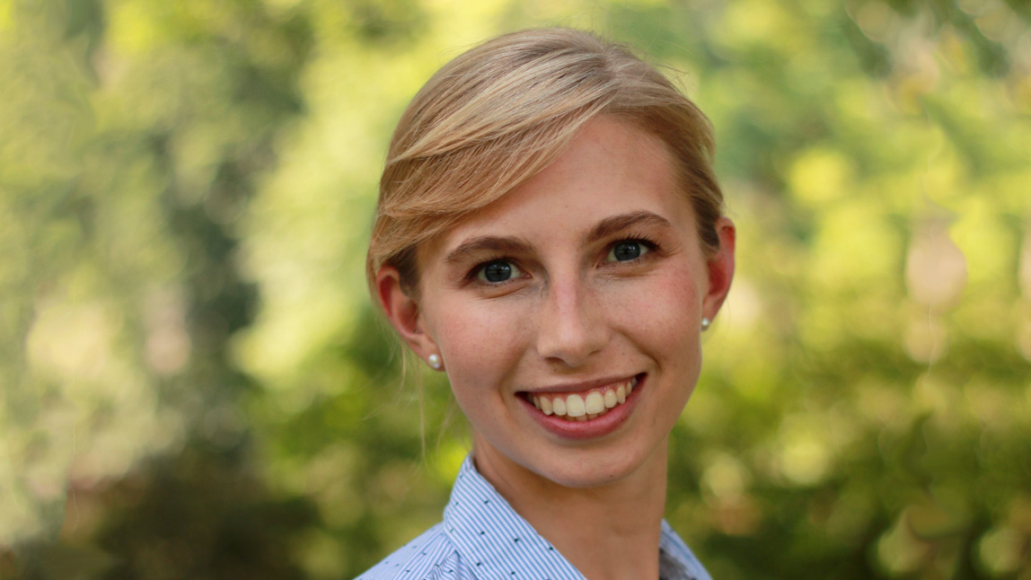 Person with blonde hair tied back in front of trees.